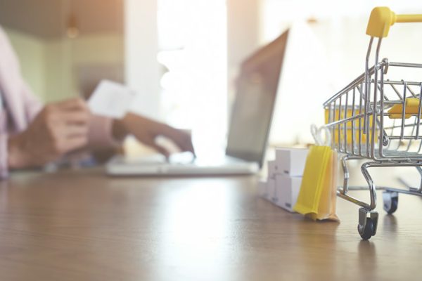 Imagem traz um carrinho de compras e uma pessoa usando um computador para ilustrar a defesa do consumidor
